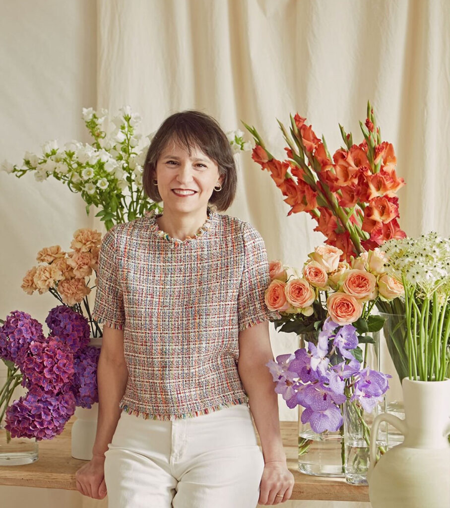 Sylvia Bustamante, founder of Madrid Flower School, sitting on a table filled with buckets of colorful flowers