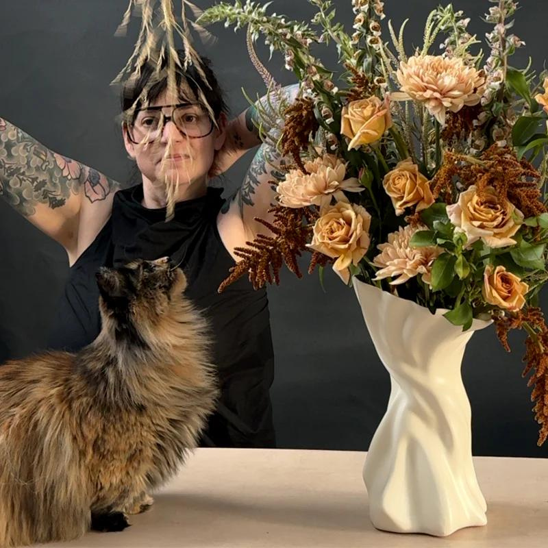 Sullivan Owen in the studio with a vase she made containing flowers in shades of caramel and butterscotch as her cat looks on